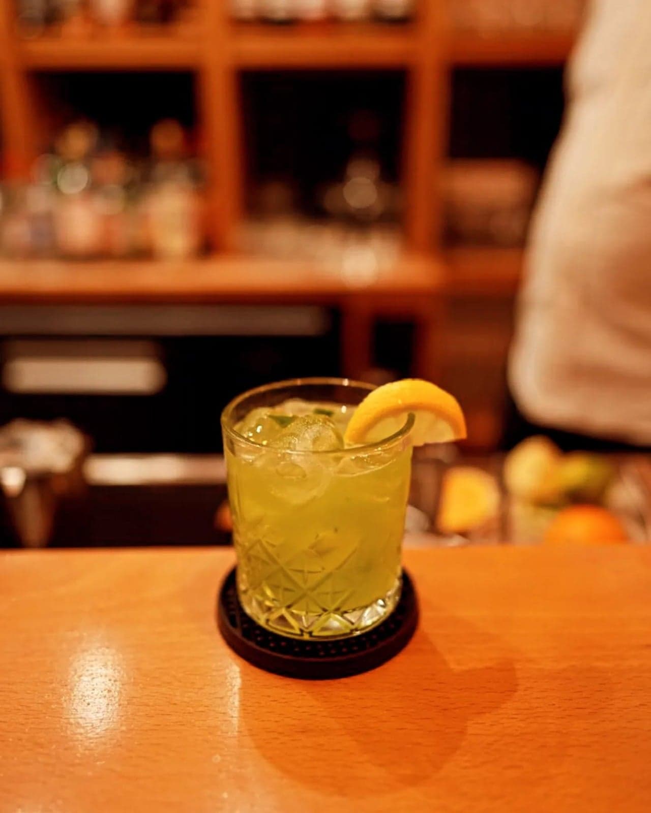 Un cocktail vert pâle avec glaçons et tranche de citron, posé sur le comptoir devant les étagères du bar